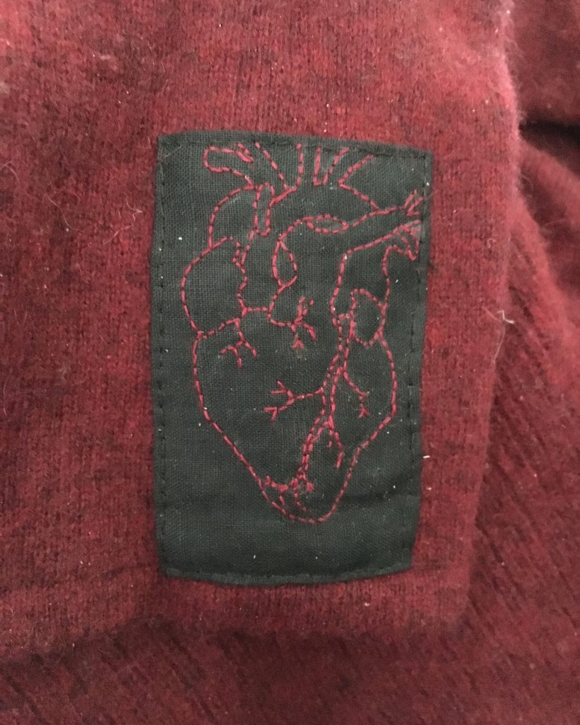 A black patch on the sleeve of a red cashmere sweater. An anatomical heart is embroidered in back stitch on the patch in red thread.
