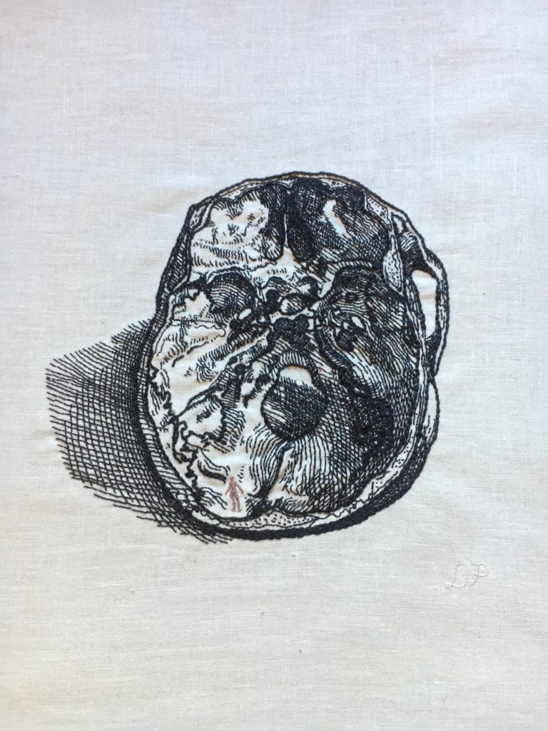 An embroidery on bone coloured linen of the inside of a human skull looking down from a cut open top done in thin black thread. The textures make it look like the Vesalius woodcut it is based on. In the bottom left corner of the skull there is the tiny silhouette of a woman done in bronze thread.