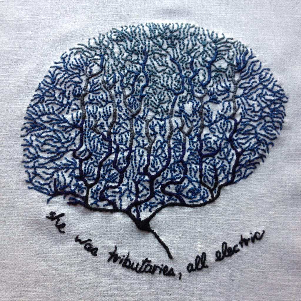 An embroidery on bone white linen. There are many coral-like neurons in the vague shape of a brain embroidered in shades of blue. The bottom section is navy blue and transitions to a lighter blue at the top. The text at the bottom reads: she was tributaries, all electric.