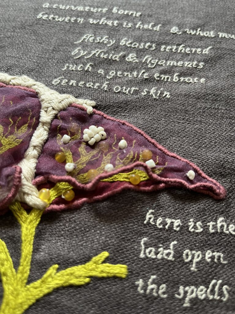 A detail shot of the small lobe on Lia's liver embroidery. There are small yellow beads on the biliary system inside the smaller lobe. There are white beads sewn on the narrow end of the top organza liver. A block of text stitched in light grey single strand thread can be read on top of the smaller lobe. It says: Fleshy beasts tethered. By fluid and ligaments. Such a gentle embrace. Beneath our skin. 