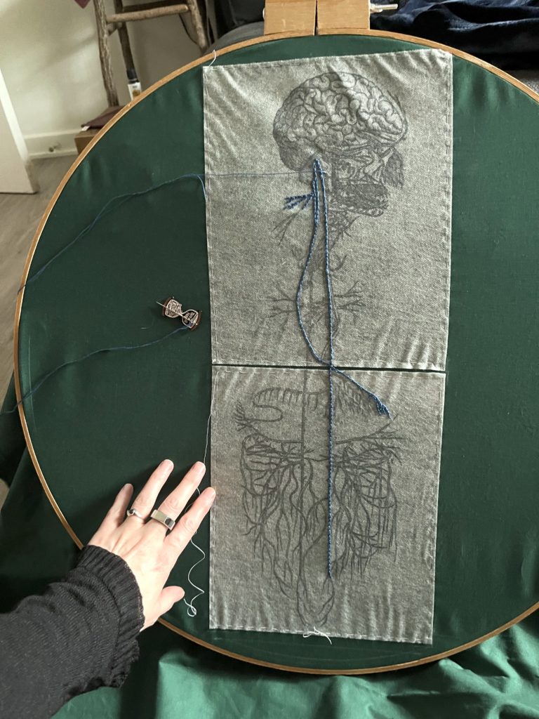 An image of an old engraving of a vagus nerve is printed on sulky material and basted to green cotton in a huge 2 foot wide wooden embroidery hoop. Some stitching of one of the nerve lines has been done in blue chain stitch in perlé cotton. Lia's hand can be seen next to the image.