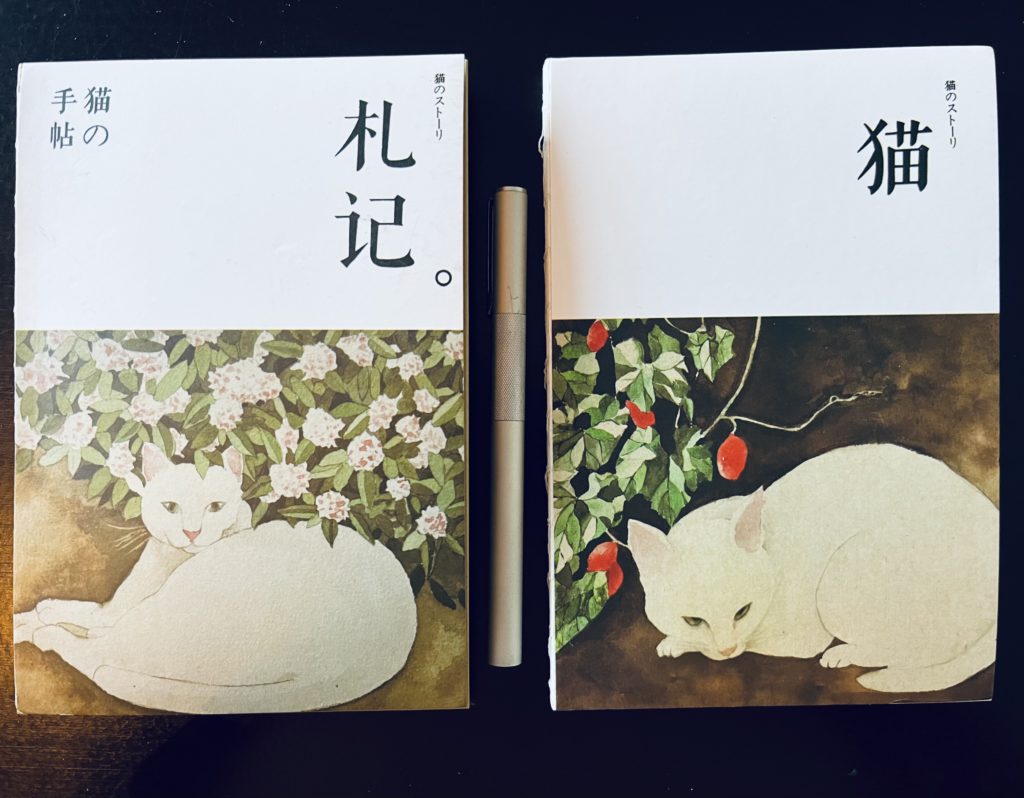 Two paperback journals on a wooden desk with a silver pen between them. Both have paintings of white cats on the bottom half of the cover and Japanese text on the top half. The left journal has a cat lounging under hydrangeas and the right has a cat curled up under a goji berry bush. 