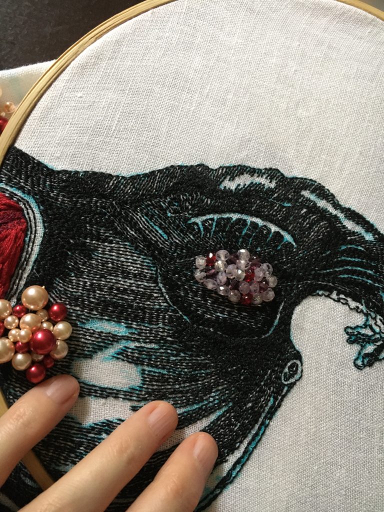 An embroidery of a uterus with the mesometrium like wings stitched in black thread on white linen looking like a woodcut. There is a strawberry sized section of red, pink, and cream beads of various sizes on the left and a strawberry sized section of clear, purple, and red beads over the ovary.