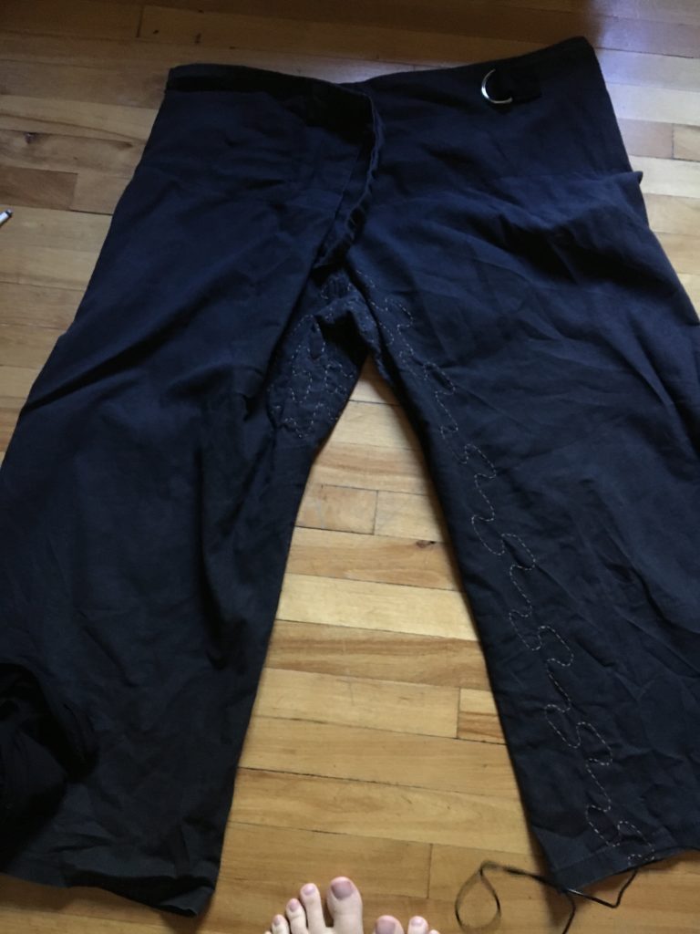 A pair of black wide-legged capri pants lying on a wooden floor. In the crotch and along the inner left leg is brown stitching in a Japanese sashiko rising steam stitch. 