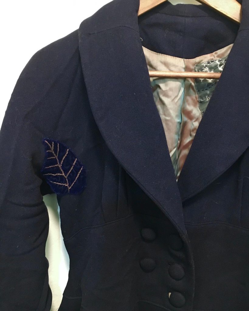 A 1940s navy blue wool coat hung against a white wall. There is a velvet leaf in the right armpit and a silver patch can be seen in the lining.