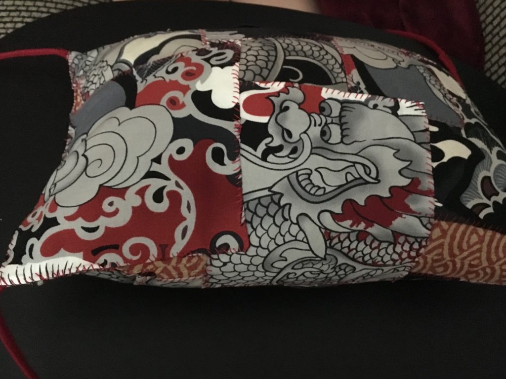 A small pillow with many patches of an Asian dragon pattern in greys, blacks, and reds, sewn on with red thread. Some of the original red and tan Japanese wave pattern cloth of the pillow can be seen on the bottom edge.