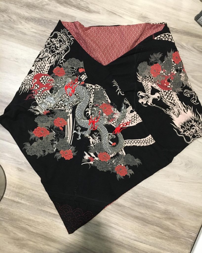 A shawl made with black t-shirt material with dragons and flowers printed and machine embroidered on it in greys, whites, and reds. The lining of the shawl is red and white seigaiha cloth. There is some red Sashiko stitching on the front point of the shawl.