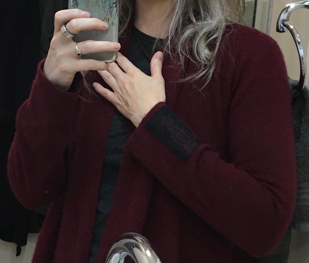 Lia in a selfie in her bathroom mirror. She is wearing a burgundy cardigan with a black patch near the cuff of the left sleeve. She has long greying hair. The bathroom is white with a cabinet and dark housecoats hung on the wall behind her.
