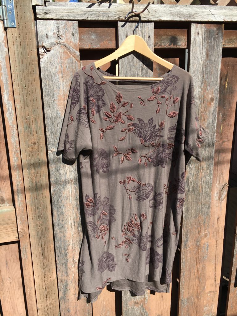 A brown-rose lightweight merino wool t-shirt dress hangs on a hanger against a fence. There are large purple peonies printed on the dress and numerous dusky rose leaves embroidered all over connected by stem stitch. 