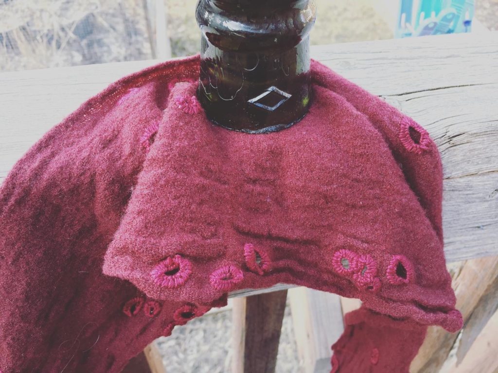 A close up of a red wool scarf with holes surrounded by red buttonhole stitch. A glass weight holds the scarf down on the edge of a deck.