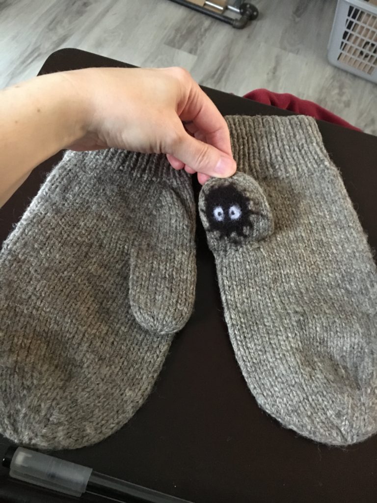 Lia’s hand holding the thumb of one of a pair of grey knitted mittens. A needle felted soot sprite with wide open eyes is on the pad of the thumb.
