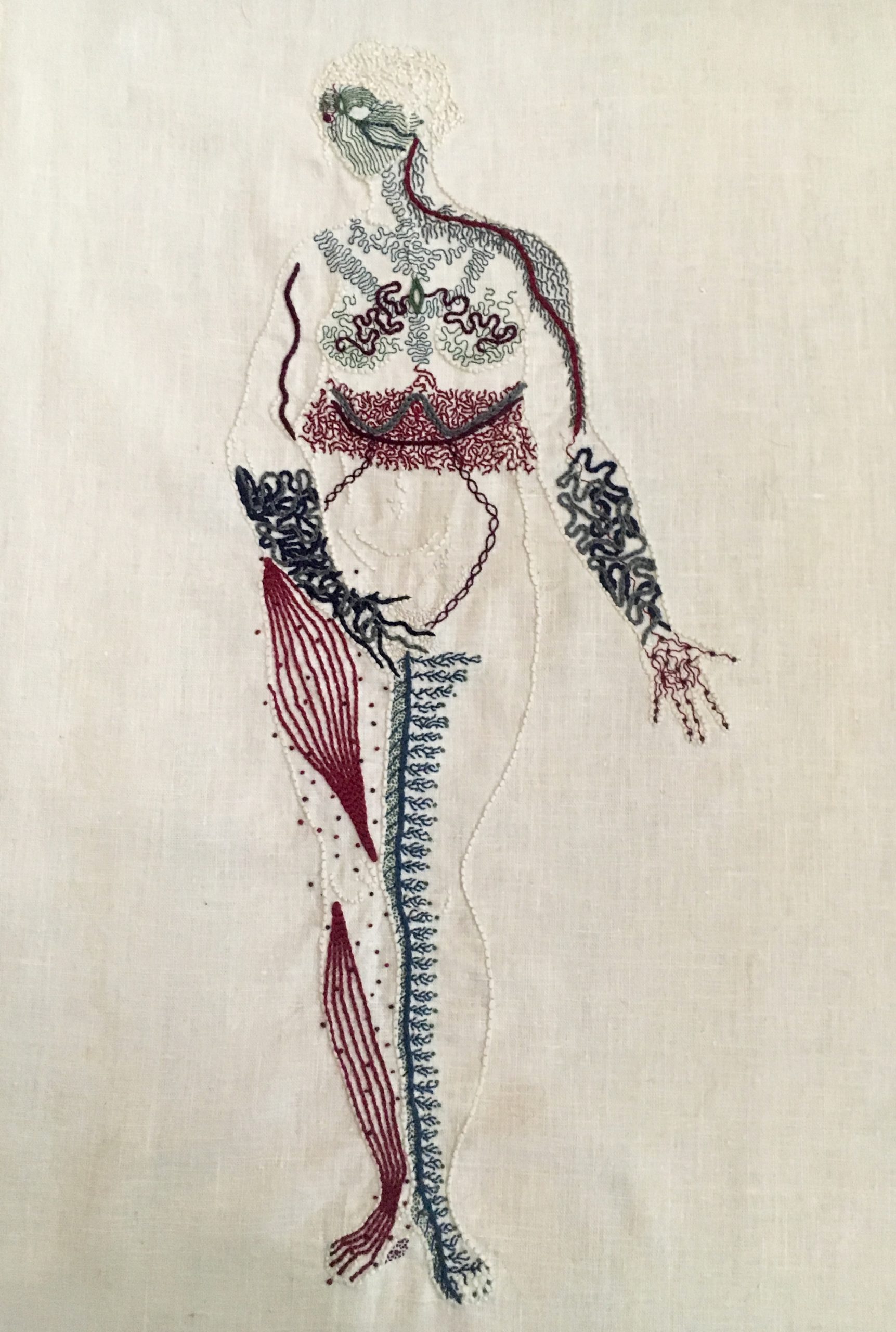 An outline of a naked woman is embroidered on linen in the same bone white colour as the linen. She stands legs together, her right hand covering her groin, her left hand, palm up, extended slightly to her side. She looks to the right. Her entire body except for her belly is covered in intricate markings representing different neurological sensations. Her face is a mask of green lines, feathery grey lines cover her shoulders and chest. There is a thick band of intricate burgundy stitching around her waist. Her forearms and hands are covered in thick blue undulant lines. Her right leg has bands of burgundy along the muscles, with small dots around them. Her inner left leg has a thick line of blue running up it, with thin branches spreading towards her outer leg.