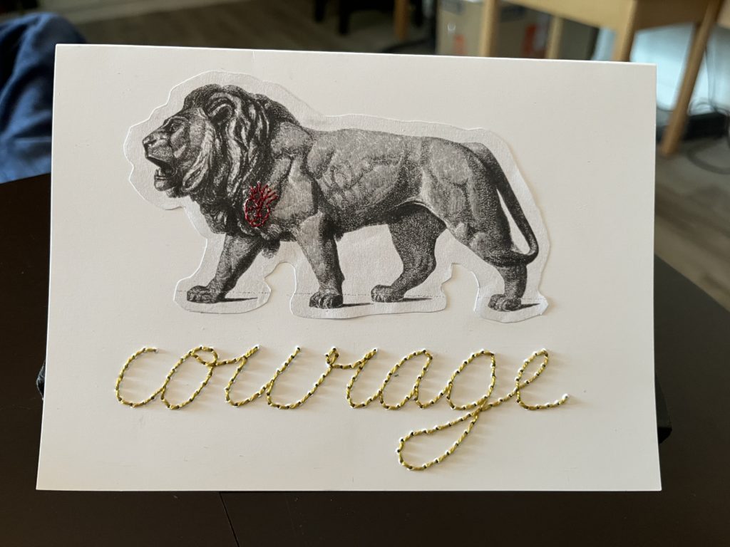 A white landscape formatted card. A black and white Victorian engraving of a lion has been printed, cut to shape, and glued on the top 2/3 of the card. There is a tiny red anatomical heart embroidered on the lion's chest. Below the lion the word "courage" is stitched in large cursive writing. 