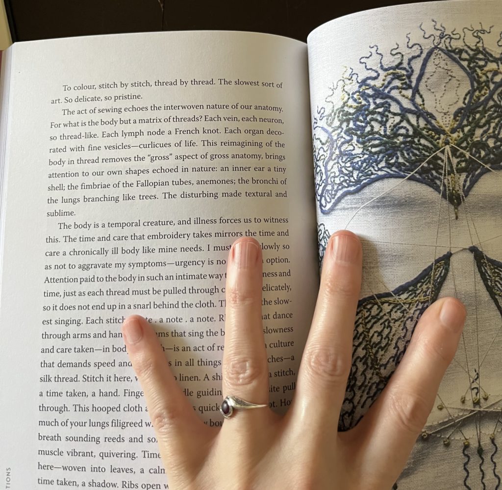 Lia’s hand holds open a spread in the Sharp Notions book with a page of her essay on the left and her paresthesia face embroidery on the right.
