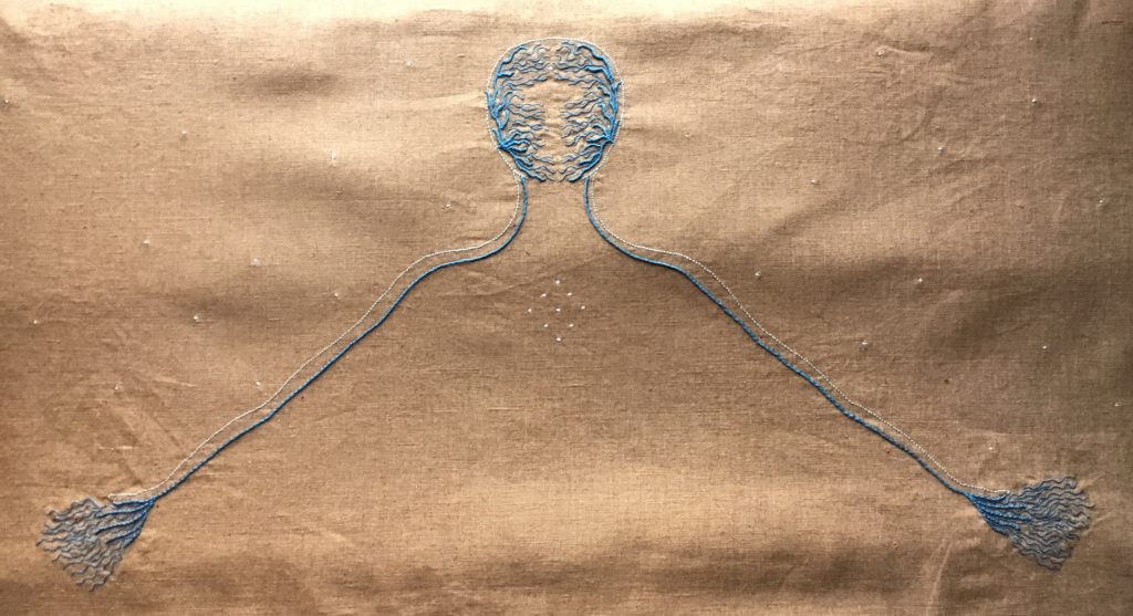 An embroidery on ochre linen, there is the outline of a head and outstretched arms. Tiny blue filigree decorates the face and hands and there are light coloured stars on the chest area and around the neck. 