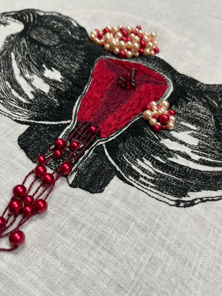 An embroidery of a uterus, including Fallopian tubes and ovaries, stitched in black thread on white linen textured like a woodcut. The photo is taken at an angle to show the textures. Inside the uterus, the endometrium is thickly stitched in bright red and burgundy. There is a large fibroid on the top of the uterus and a smaller fibroid on the right lower side, both beaded in red, pink, and cream beads. A cyst on the right ovary is beaded in clear, purple, and red beads, and a small section of dark red and purple beads is a polyp inside the uterus. Long burgundy strings hang from the bottom of the uterus with red beads on them that extend past the vaginal opening. 