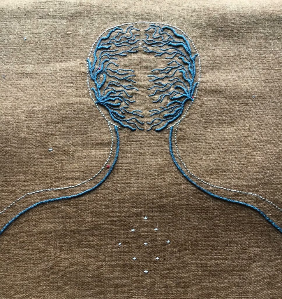 An embroidery on ochre linen. There is the outline of a head and neck. There are blue filigree lines coming from the sides of the head outline towards the centre of the face. A fine off white line outlines the head and moves down the neck and out along the arms. On the centre chest is a cluster of off-white crosses and a few sparse crosses are around the head and shoulders as well. 
