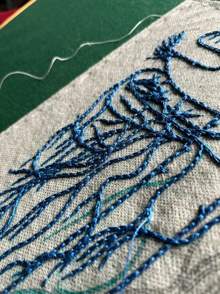 A close up of an embroidery focused on the left side of the abdominal vagus stitched on sulky material over dark green cotton. There are many layers of crossing wavy lines done in bright blue chain stitch.
