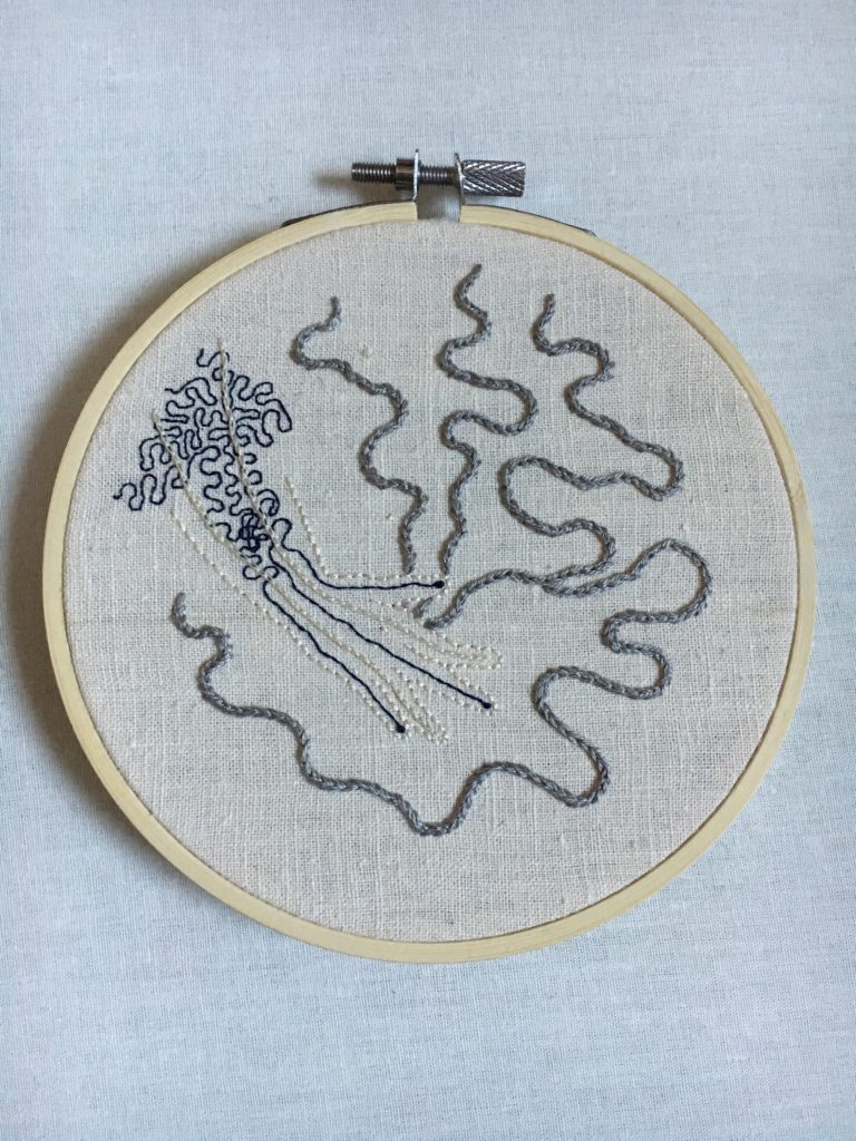 In a small embroidery hoop on bone coloured linen is the outline of a hand done in the same colour. Emanating from the palm of the hand are thick grey chain stitched lines that curl around the hand. On the back of the hand and up the lower arm are fine navy blue curls that get more dense as they move up the arm. 