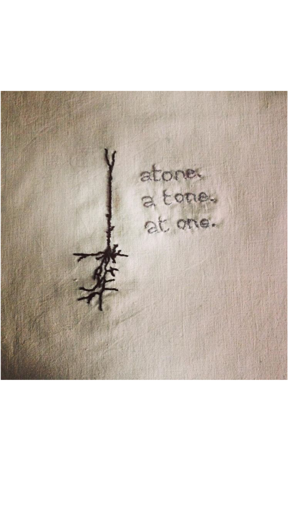 On bone white linen there is a pyramidal neuron stitched in black thread. To the right of it are three lines of text embroidered in grey that read: Atone. A tone. At one. 