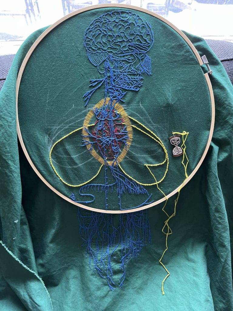 An image of an old engraving of a vagus nerve is embroidered on green cotton. The head and chest are in a large bamboo embroidery hoop. The embroidery of the main nerve lines is done in blue chain stitch in perlé cotton. The brain and some of the face is stitched in the same thread in backstitch. Details in the face are stitched in single strand floss. A bright red anatomical heart is stitched in two and one strand red whipped back stitch. Around it a bright golden yellow halo in a vesica piscis shape is stitched in single strand thread. Three sets of ribs have been sketched in chalk and the lowest and second lowest ribs are being stitched in bright green/yellow stem stitch. A needle minder in the shape of an hourglass that reads, "This is taking forever," sits near the stitches holding a needle. 