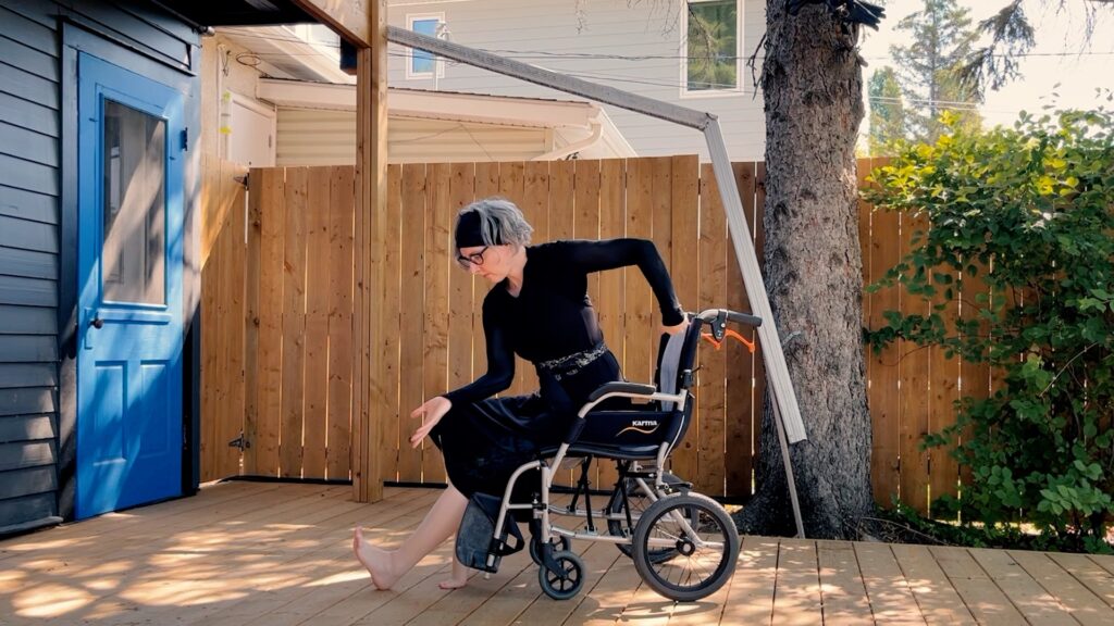 Lia, a white woman with a short grey bob held back in a headband wears a black dress and sits in a wheelchair. Her arms are in angular positions and he left leg is stretched out. Behind her is a wooden fence and a bright blue door is to the left of the image.